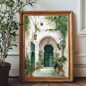 May include: A framed watercolour print shows an arched doorway with a green door, surrounded by greenery and potted plants. The artwork is set in a gold frame and rests on a wooden surface, creating a serene, inviting scene.
