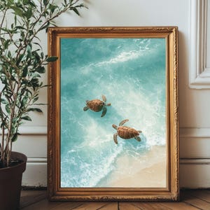 May include: A framed art print depicting two sea turtles in turquoise water near a sandy beach. The artwork is presented in a gold-coloured ornate frame, enhancing the coastal theme. The print is set against a white wall and wooden floor.