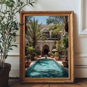 May include: A framed print of a courtyard with a turquoise pool, palm trees, and ornate architecture. The gold frame rests on a wooden floor, with a potted plant visible on the left.