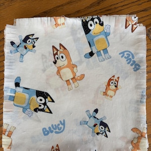 May include: White fabric square featuring cartoon characters in blue, orange, and yellow. The word "Bluey" is printed in blue. The fabric is likely intended for crafting or sewing projects.