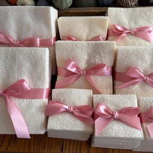 May include: A collection of square, off-white felt stacks of varying sizes, each neatly tied with a smooth, shiny pink satin ribbon. The ribbons are tied into bows at the center of each stack, creating a uniform and organized presentation of the textile squares.