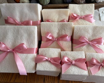 May include: A collection of square, off-white felt stacks of varying sizes, each neatly tied with a smooth, shiny pink satin ribbon. The ribbons are tied into bows at the center of each stack, creating a uniform and organized presentation of the textile squares.