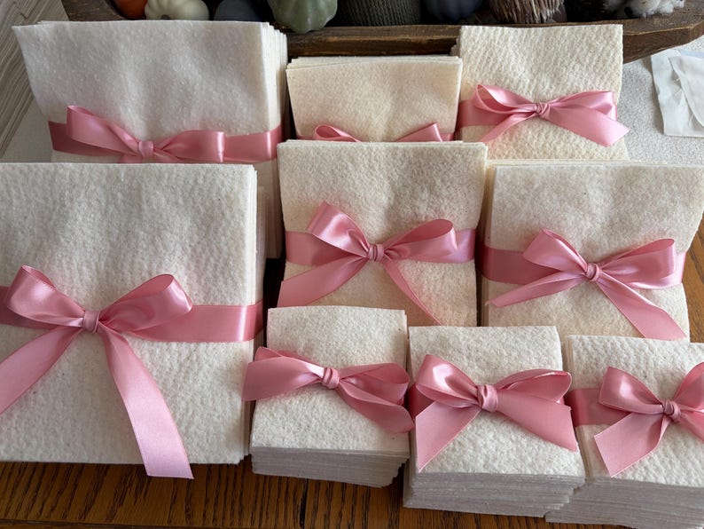 May include: A collection of square, off-white felt stacks of varying sizes, each neatly tied with a smooth, shiny pink satin ribbon. The ribbons are tied into bows at the center of each stack, creating a uniform and organized presentation of the textile squares.