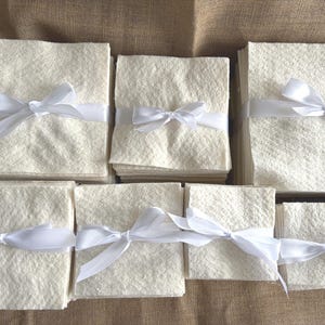 May include: Several stacks of off-white, textured paper squares are arranged on a neutral surface. Each stack is tied with a white satin ribbon, forming a bow. The paper has a subtle, organic texture, suggesting a handmade or natural quality.