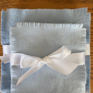 May include: A stack of light blue fabric squares with fringed edges, tied with a white satin ribbon bow. The fabric has a soft texture and is neatly arranged, creating a simple, elegant presentation.