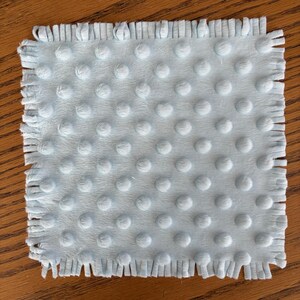 May include: A square, light blue baby blanket with a textured, dotted surface and a fringed edge. The blanket is made of a soft material and is likely used for swaddling or as a comfort item for infants.