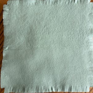 May include: A square piece of light sage green fabric with a fringed edge. The fabric has a soft, matte texture and features evenly spaced vertical cuts along all four sides, creating a uniform fringe border around the central square panel.