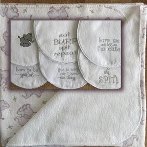 May include: A white baby blanket with a gray elephant pattern and a set of white burp cloths featuring gray embroidered text including "eat BURP spit repeat," "born to be spoiled," "this is why I can't wear nice things," "burp me we tell me I'm cute," and "oh SPIT!"