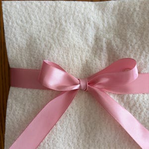 May include: A square, off-white, textured felt material folded or layered, wrapped horizontally with a smooth, shiny, light pink satin ribbon tied into a centered bow with two long, angled tails extending downward.