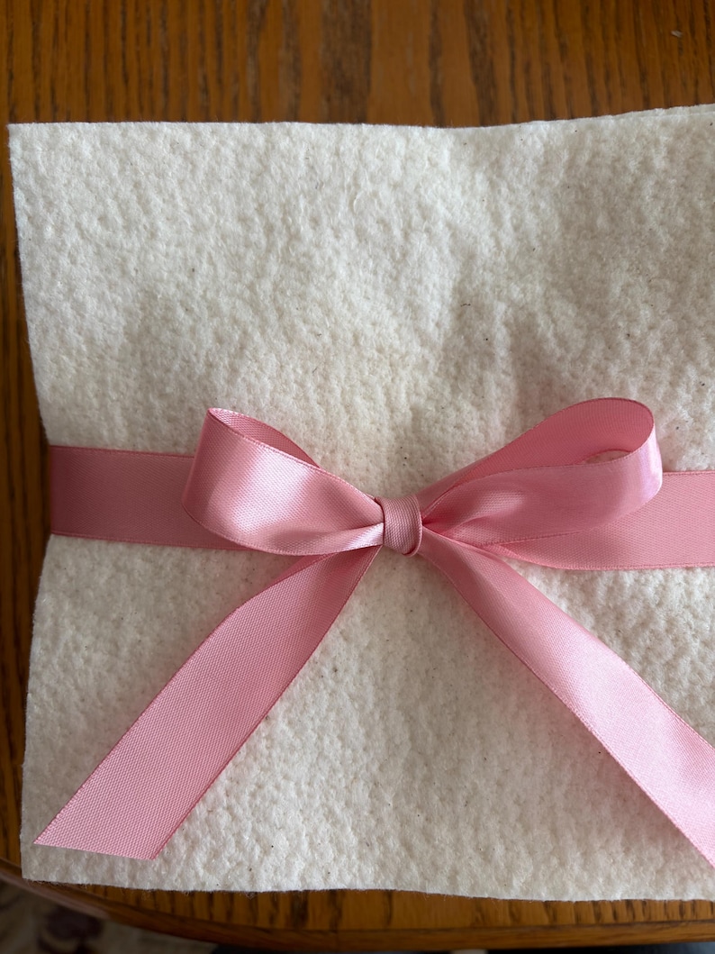 May include: A square, off-white, textured felt material folded or layered, wrapped horizontally with a smooth, shiny, light pink satin ribbon tied into a centered bow with two long, angled tails extending downward.