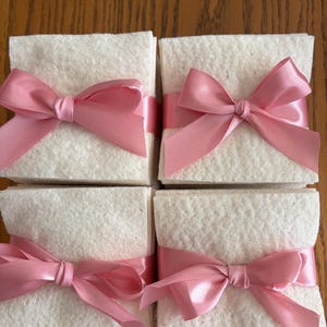 May include: Four square stacks of off-white, textured paper sheets, each neatly bundled with a smooth, shiny pink satin ribbon tied into a decorative bow at the front center. The stacks are arranged in a two-by-two grid formation.