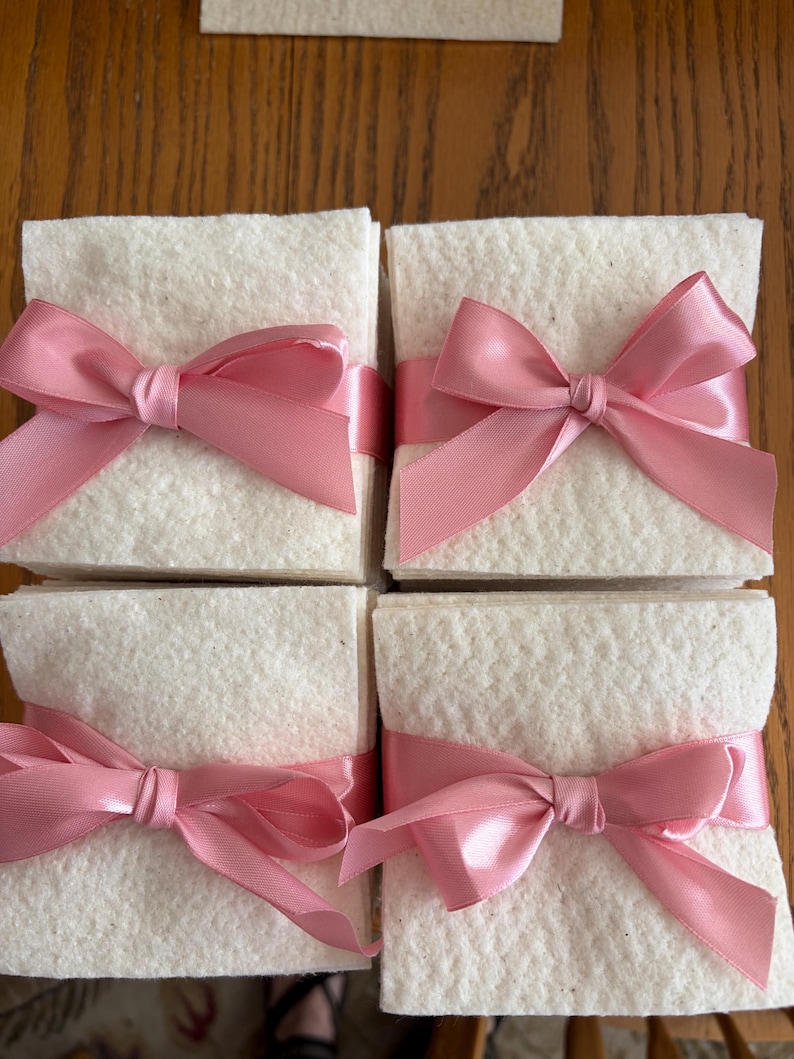 May include: Four square stacks of off-white, textured paper sheets, each neatly bundled with a smooth, shiny pink satin ribbon tied into a decorative bow at the front center. The stacks are arranged in a two-by-two grid formation.