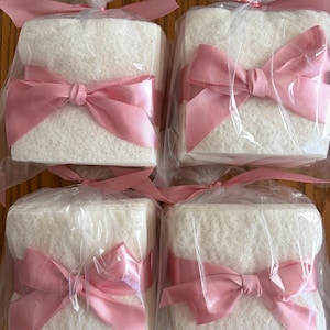 May include: Four square stacks of white textured paper napkins, each individually wrapped in clear plastic packaging and tied with a decorative pink satin ribbon bow at the front, arranged in a two-by-two grid formation.