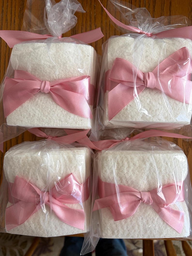 May include: Four square stacks of white textured paper napkins, each individually wrapped in clear plastic packaging and tied with a decorative pink satin ribbon bow at the front, arranged in a two-by-two grid formation.