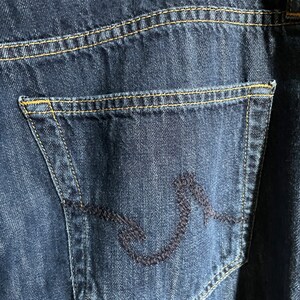 May include: Close-up of dark blue denim jeans with a back pocket featuring a decorative stitched design. The jeans have a brown leather patch with a logo and yellow stitching.