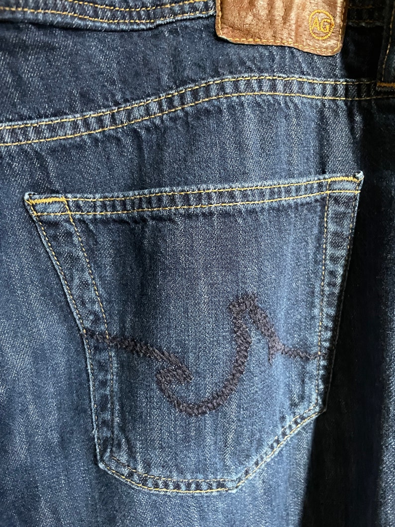 May include: Close-up of dark blue denim jeans with a back pocket featuring a decorative stitched design. The jeans have a brown leather patch with a logo and yellow stitching.