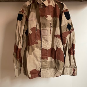 Könnte beinhalten: Eine Tarnjacke mit Knopfleiste und zwei Reißverschlusstaschen. Die Jacke hat schwarze Patches an den Ärmeln.