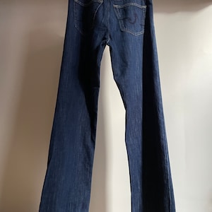 May include: Dark wash denim jeans hanging on a wooden hanger. The jeans have a classic five-pocket design, a button and zipper closure, and a leather patch on the back. The jeans are a deep indigo color.