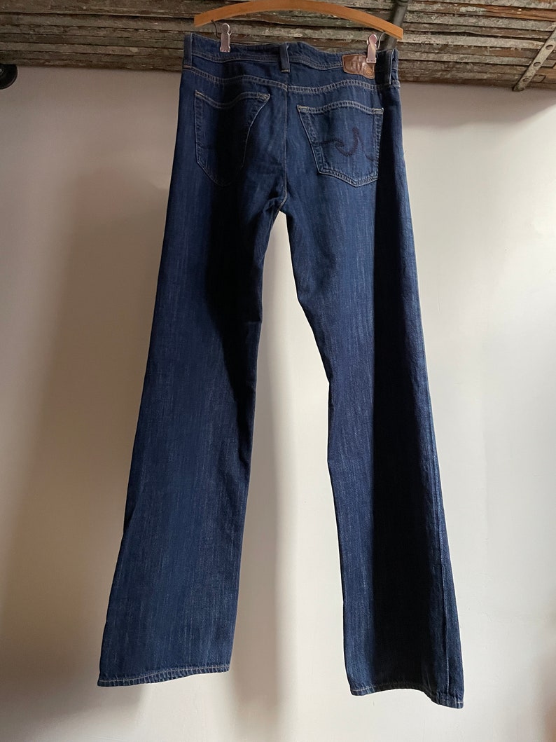 May include: Dark wash denim jeans hanging on a wooden hanger. The jeans have a classic five-pocket design, a button and zipper closure, and a leather patch on the back. The jeans are a deep indigo color.