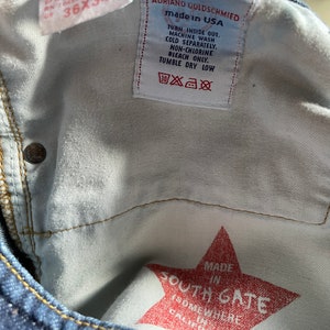 May include: Close-up of the inside of blue denim jeans. The labels read "ADRIANO GOLDSCHMIED made in USA" and "36x34". A red star-shaped graphic reads "MADE IN SOUTH GATE (SOMEWHERE IN CALIFORNIA)".