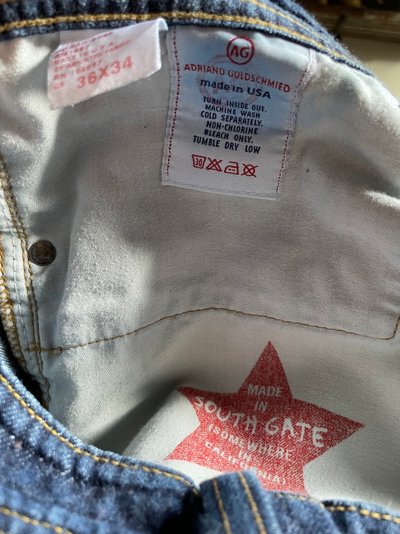 May include: Close-up of the inside of blue denim jeans. The labels read "ADRIANO GOLDSCHMIED made in USA" and "36x34". A red star-shaped graphic reads "MADE IN SOUTH GATE (SOMEWHERE IN CALIFORNIA)".