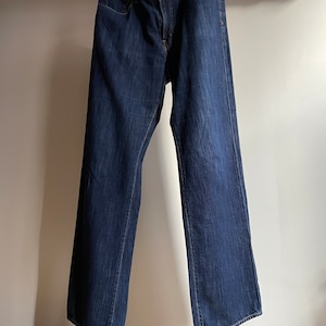 May include: Dark wash denim jeans hanging on a wooden hanger. The jeans have a classic five-pocket design, a button and zipper closure, and a straight leg fit. The jeans are a medium weight and appear to be pre-worn.