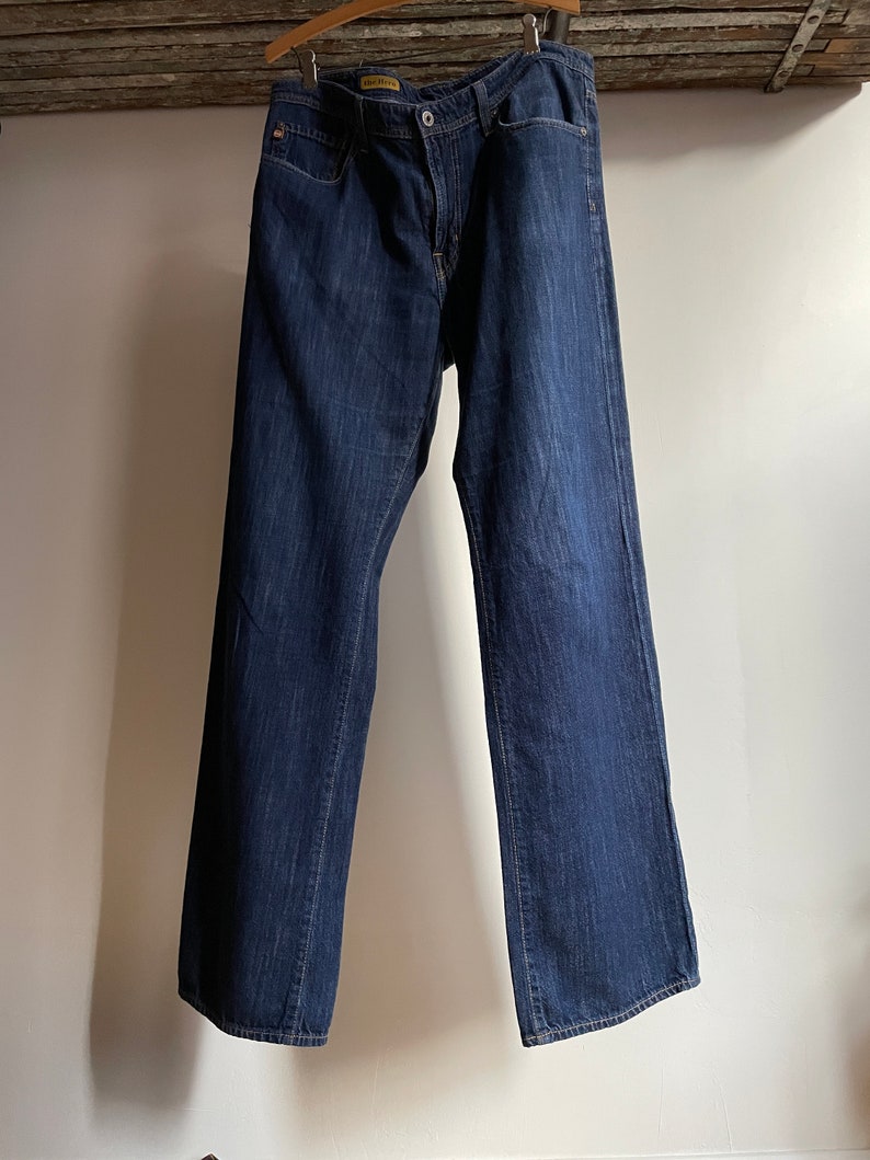 May include: Dark wash denim jeans hanging on a wooden hanger. The jeans have a classic five-pocket design, a button and zipper closure, and a straight leg fit. The jeans are a medium weight and appear to be pre-worn.