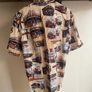 May include: Short-sleeved button-down shirt with a cream-colored background and a repeating pattern of vintage city scenes in shades of brown, red, and blue. The shirt has a classic collar and is hanging from a wooden rod.