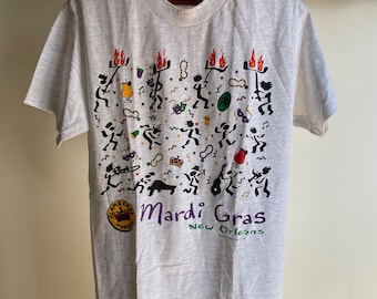 May include: A light gray t-shirt with a colorful Mardi Gras design. The graphic features stick figures, musical instruments, and the words "Mardi Gras New Orleans". The shirt is hanging on a wooden hanger.