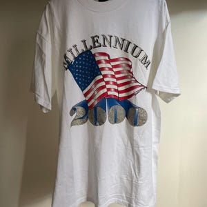 Op de afbeelding: Wit T-shirt met een grafisch ontwerp. Het ontwerp toont het woord "MILLENNIUM" boven een wapperende Amerikaanse vlag en het nummer "2000". Het ontwerp bevat rode, witte en blauwe kleuren, met een zilverkleurig lettertype en accenten.