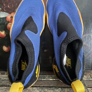 May include: A pair of blue and black Nike water shoes. The shoes have a mesh upper, a black collar, and a yellow pull tab. The Nike logo is visible on the side and insole. The soles are a light brown color.