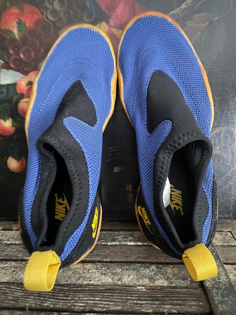 May include: A pair of blue and black Nike water shoes. The shoes have a mesh upper, a black collar, and a yellow pull tab. The Nike logo is visible on the side and insole. The soles are a light brown color.
