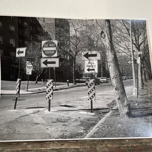 Può includere: Una fotografia in bianco e nero di un incrocio con diversi segnali stradali. I segnali indicano "Divieto di ingresso", "Tutto il traffico" e frecce direzionali. I segnali sono montati su pali e sono visibili sullo sfondo di alberi ed edifici.