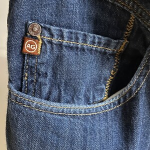 May include: Close-up of a dark blue denim pocket with yellow stitching. A small brown tag with the letters "AG" is attached to the pocket. A silver button is visible above the tag.