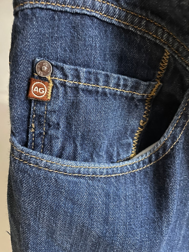 May include: Close-up of a dark blue denim pocket with yellow stitching. A small brown tag with the letters "AG" is attached to the pocket. A silver button is visible above the tag.