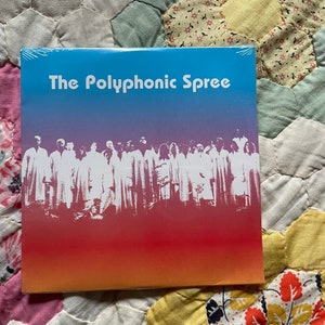 Op de afbeelding: Een cd-hoesje met de witte tekst "The Polyphonic Spree". De achtergrond loopt over van blauw naar rood, met een wit silhouet van een groep mensen. De cd ligt op een bloemenquilt.