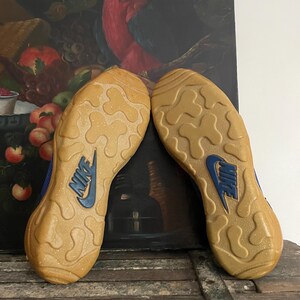 May include: Pair of Nike sneakers, soles up, showing a tan rubber outsole with a blue Nike logo. The shoes have a blue and tan color scheme. The background features a painting of a parrot and fruit.