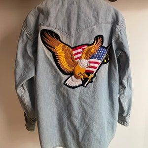 Può includere: Una camicia di jeans blu chiaro con una grande toppa ricamata sul retro. La toppa presenta un'aquila dorata con le ali spiegate che tiene una bandiera americana.