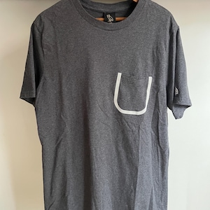 Può includere: Una maglietta grigio antracite con tasca bordata di bianco. La maglietta ha un girocollo e maniche corte. Il logo del gufo OVO è visibile sulla maglietta e sulla manica.