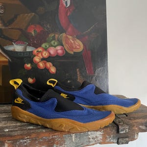 May include: A pair of blue and black Nike water shoes with a brown sole. The shoes have a slip-on design with a yellow pull tab and the Nike logo. The shoes are on a wooden surface.