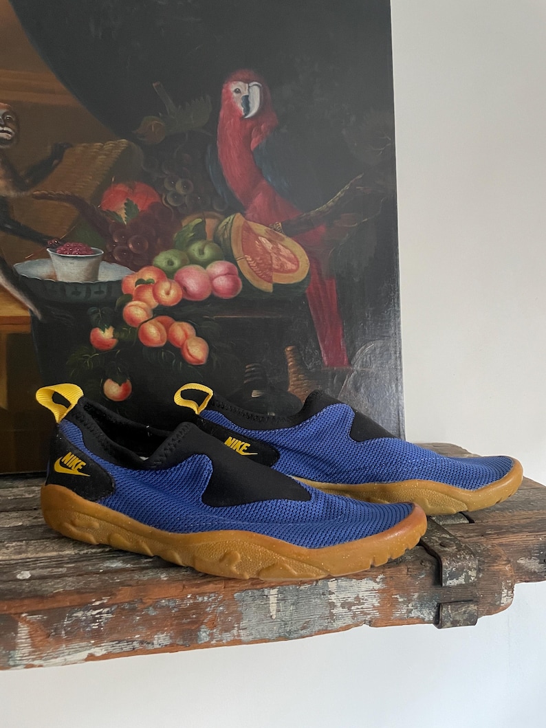 May include: A pair of blue and black Nike water shoes with a brown sole. The shoes have a slip-on design with a yellow pull tab and the Nike logo. The shoes are on a wooden surface.