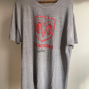 May include: A gray t-shirt featuring a red Dodge logo with a ram's head inside a shield. The word "DODGE" is above the logo, and "EVERNHAM" is printed below. The shirt is hanging on a wooden hanger.