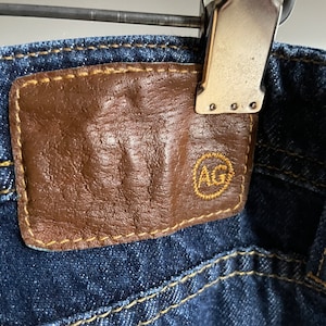 May include: Close-up of dark wash denim jeans with a brown leather patch. The patch has the letters "AG" in a gold-colored circle. The jeans are held by a metal clip.