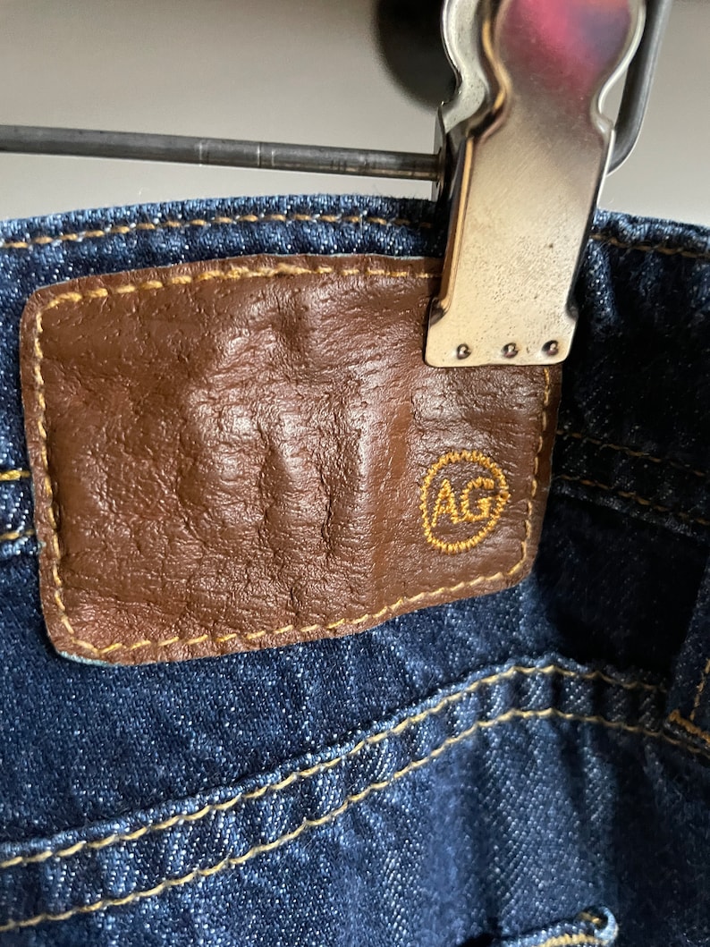 May include: Close-up of dark wash denim jeans with a brown leather patch. The patch has the letters "AG" in a gold-colored circle. The jeans are held by a metal clip.