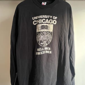 Puede incluir: Camiseta negra de manga larga con el texto "UNIVERSITY OF CHICAGO" y un escudo. El escudo presenta un águila y las palabras "HELL DOES FREEZE OVER". La camiseta está colgada en una percha de madera.