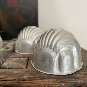 May include: Three vintage, silver-colored metal molds with a fluted, dome-like design. The molds are sitting on a weathered, dark wooden surface. The background includes a painting with dark and red tones.