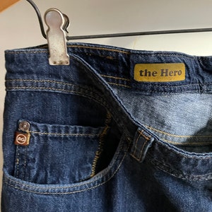 May include: Close-up of dark blue denim jeans with visible stitching and a small pocket. The jeans feature a "the Hero" label and a small logo tag. The jeans are held by a metal clip on a wooden hanger.
