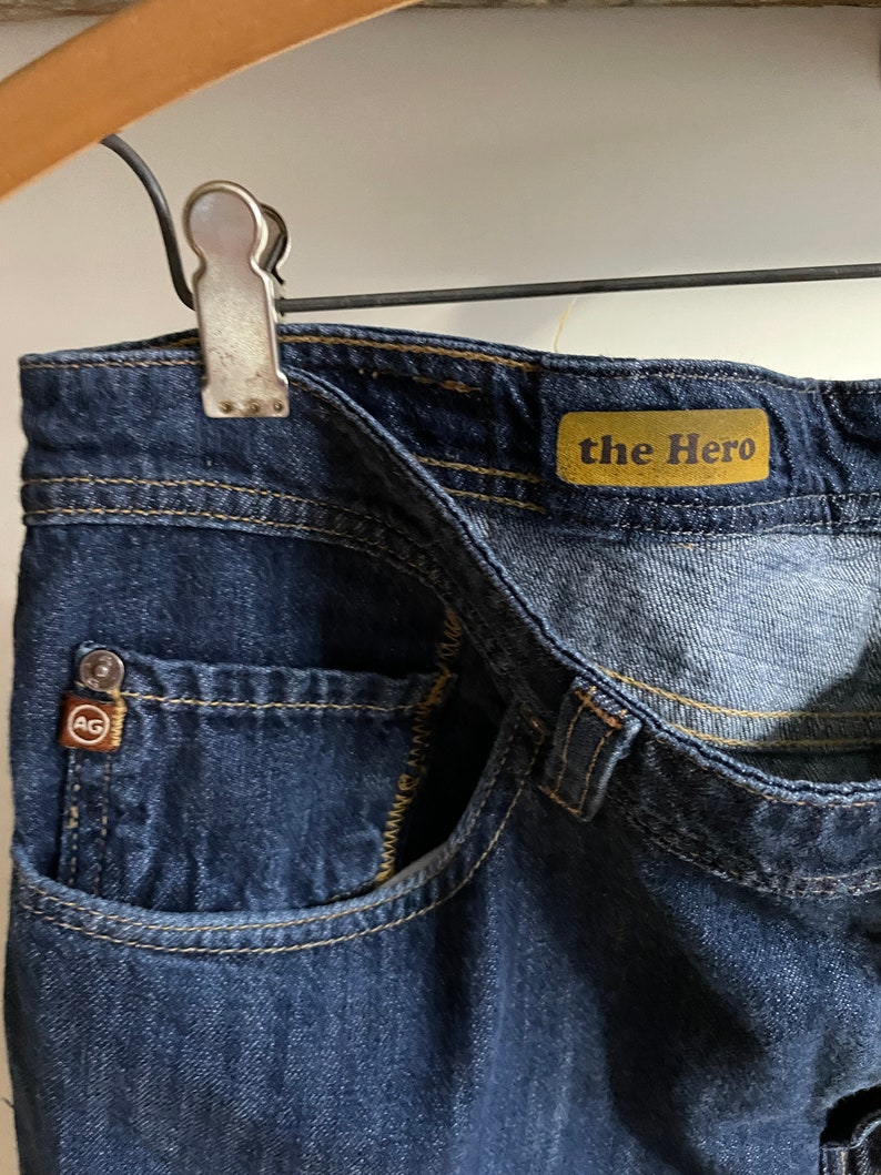 May include: Close-up of dark blue denim jeans with visible stitching and a small pocket. The jeans feature a "the Hero" label and a small logo tag. The jeans are held by a metal clip on a wooden hanger.