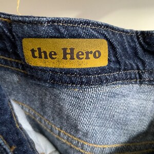 May include: Close-up of the waistband of dark blue denim jeans. A mustard-colored tag is sewn onto the jeans, displaying the text "the Hero" in a dark brown font. The stitching is a contrasting yellow color.