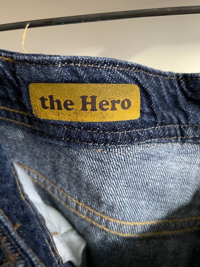May include: Close-up of the waistband of dark blue denim jeans. A mustard-colored tag is sewn onto the jeans, displaying the text "the Hero" in a dark brown font. The stitching is a contrasting yellow color.